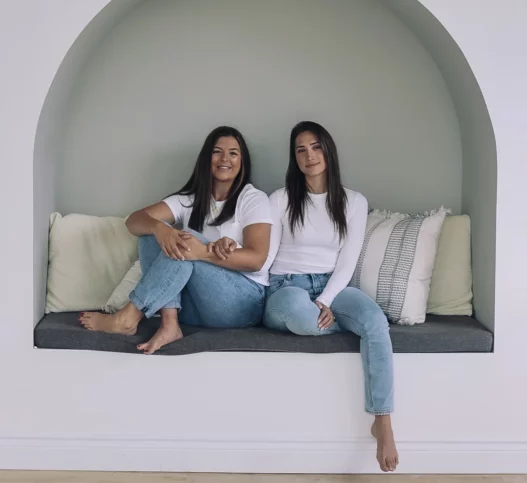 Pelvic health professionals Laura and Nadia sitting in a nook smiling while wearing a white shirt and blue jeans